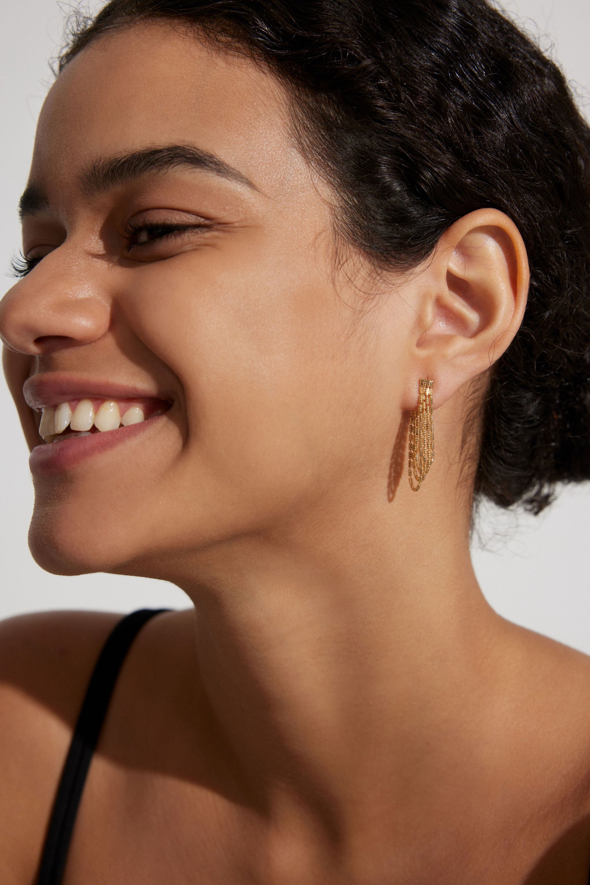 A person wearing a smile on their face, showcasing a pair of gold-colored tassel earrings with silver hooks.
